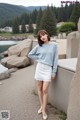 a woman standing in front of a lake wearing a blue sweater and white skirt